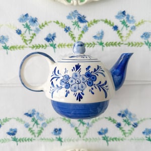 May include: A blue and white teapot with a floral design. The teapot is sitting on a white tablecloth with blue and green embroidered flowers.