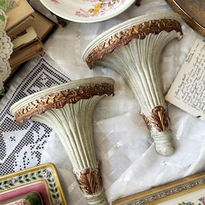 May include: Two wall-mounted shelves featuring a fluted, fan-shaped design in a distressed off-white finish. The curved top edges and the base of each shelf are accented with ornate, raised gold-toned detailing, providing a textured appearance.