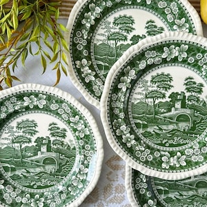 May include: A collection of green and white ceramic plates with a landscape design. The plates feature a scalloped edge with floral detailing. The central design depicts a bridge, trees, and a building in a scenic setting.