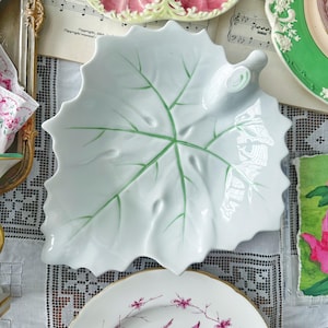 May include: White ceramic leaf-shaped serving dish with green vein detailing. The dish is surrounded by other decorative plates with floral and shell designs. The overall theme is vintage tableware.
