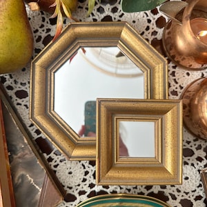 May include: Two decorative mirrors with ornate, antique-style gold frames. One mirror is octagonal in shape, while the smaller mirror is square. Both frames feature a textured, weathered gold finish with dark brown accents along the inner edges.