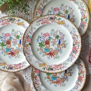 May include: A set of four white ceramic plates featuring a central floral motif with pink, blue, and green flowers, accented by brown geometric lattice patterns and a gold-trimmed scalloped edge. The plates display a repeating botanical design.