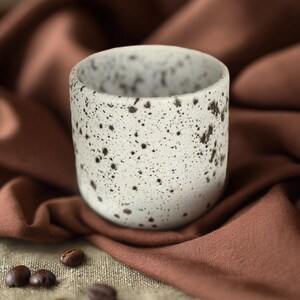 Op de afbeelding: Een gespikkelde keramische beker met een witte basis en donkerbruine vlekken. De beker staat op een gevouwen bruine doek, met verspreide koffiebonen in de buurt. Het ontwerp van de beker is minimalistisch en rustiek.