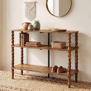 May include: A three-tier wooden console table featuring four turned, spool-style legs and a central black metal support beam. The shelves hold various items including books, a woven basket, a wooden bowl, and two small potted plants.