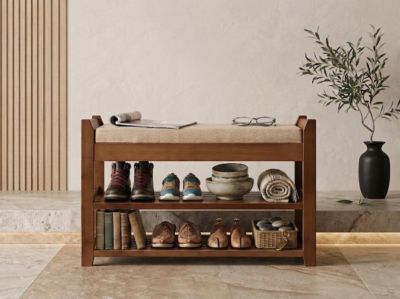Peut inclure: Un banc &agrave; chaussures en bois avec une assise rembourr&eacute;e beige, dot&eacute; de deux &eacute;tag&egrave;res de rangement &agrave; lattes. Les &eacute;tag&egrave;res contiennent divers objets dont des bottes, des baskets, des bols en c&eacute;ramique, un textile roul&eacute;, des livres et des chaussures.