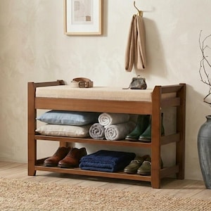 Peut inclure: Un banc d'entr&eacute;e en bois avec un coussin d'assise beige rembourr&eacute;, dot&eacute; de deux &eacute;tag&egrave;res de rangement ouvertes. Les &eacute;tag&egrave;res contiennent deux coussins, trois serviettes roul&eacute;es, une couverture bleue pli&eacute;e et quatre paires de chaussures.