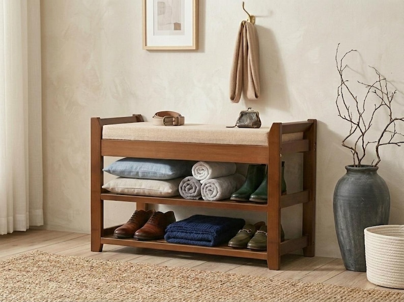 Peut inclure: Un banc d'entr&eacute;e en bois avec un coussin d'assise beige rembourr&eacute;, dot&eacute; de deux &eacute;tag&egrave;res de rangement ouvertes. Les &eacute;tag&egrave;res contiennent deux coussins, trois serviettes roul&eacute;es, une couverture bleue pli&eacute;e et quatre paires de chaussures.