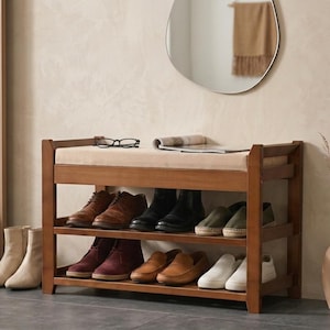 Peut inclure: Un banc &agrave; chaussures en bois avec un coussin d'assise beige rembourr&eacute;. Le meuble dispose de deux &eacute;tag&egrave;res ouvertes contenant diverses paires de chaussures, dont des mocassins en cuir marron, des bottes noires et des baskets blanches.