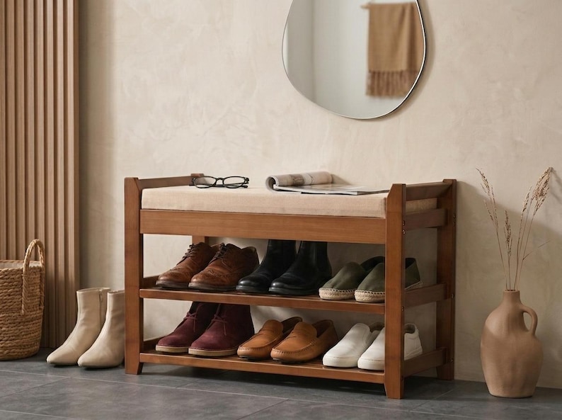 Peut inclure: Un banc &agrave; chaussures en bois avec un coussin d'assise beige rembourr&eacute;. Le meuble dispose de deux &eacute;tag&egrave;res ouvertes contenant diverses paires de chaussures, dont des mocassins en cuir marron, des bottes noires et des baskets blanches.