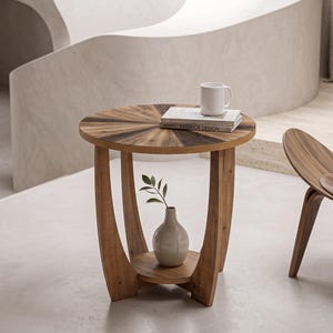 May include: A round wooden side table with a sunburst grain pattern on the top surface, featuring three curved legs and a lower circular shelf. A white ceramic mug sits on two stacked books on the table, and a small vase holds a green sprig.