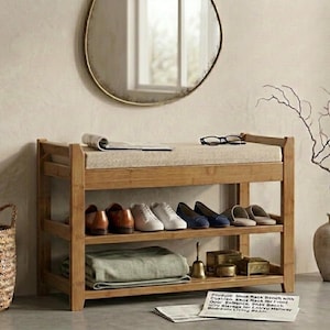 Peut inclure: Un banc &agrave; chaussures en bambou avec un coussin d'assise beige. Le meuble dispose de deux &eacute;tag&egrave;res ouvertes de rangement, contenant actuellement plusieurs paires de chaussures, un textile vert pli&eacute; et de petits objets d&eacute;coratifs.