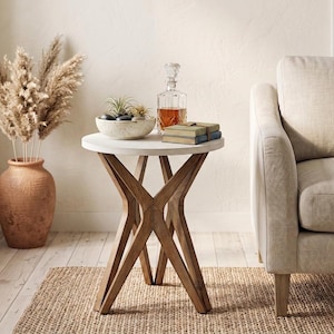 May include: A round side table with a white top and a brown wooden base featuring four intersecting legs. The table holds a speckled bowl with small plants and stones, a clear glass decanter filled with amber liquid, and two stacked books.