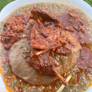 May include: A close-up shot of a plate of food featuring a large portion of a brown, doughy substance, surrounded by a green and red sauce. The dish is topped with pieces of cooked meat and a small amount of red sauce. A gold-colored utensil is visible.