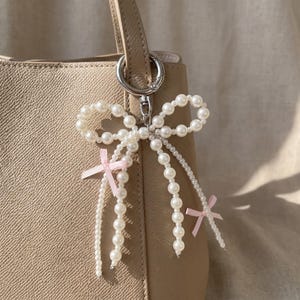 May include: A pearl beaded bow-shaped keychain with two small pink ribbons. The keychain is attached to a beige handbag with a silver-colored clasp. The bow is made of white faux pearls.