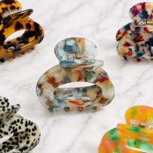 May include: A collection of colorful hair claw clips in various patterns. The clips feature a marbled design with shades of blue, orange, and cream. Each clip has a gold-colored metal clasp.