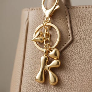 Puede incluir: Un llavero dorado con un dije de la letra "K", dos dijes de corazón y un anillo redondo. El llavero está sujeto a un bolso de cuero beige. El metal tiene un acabado brillante.