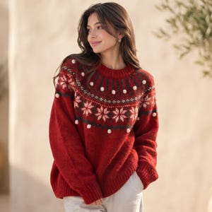 Könnte beinhalten: Ein roter, langärmeliger Strickpullover mit Rundhalsausschnitt und einer dekorativen Passe mit weißem und dunkelgrünem Schneeflockenmuster. Der Pullover ist mit kleinen weißen Bommeln verziert, die kreisförmig auf der Brust angeordnet sind.