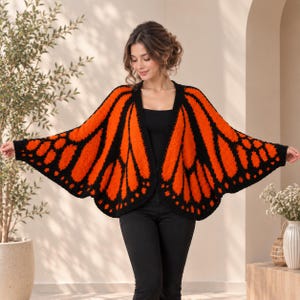 Könnte beinhalten: Eine Häkelstrickjacke mit einem leuchtend orangefarbenen und schwarzen Monarchfalter-Flügelmuster. Das Kleidungsstück hat ein offenes Design mit weiten, flügelartigen Ärmeln und einem schwarzen Rand, getragen über einem schwarzen Oberteil und einer schwarzen Hose.