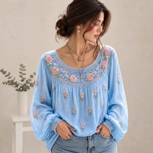 May include: A light blue long-sleeved blouse featuring a round neckline with intricate floral embroidery in shades of pink and white. The garment has gathered cuffs and a loose, flowing silhouette, decorated with small scattered floral motifs.