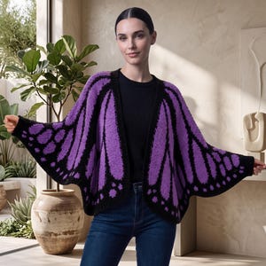 Puede incluir: Un cárdigan abierto de color morado y negro con un patrón de alas de mariposa. La prenda tiene una textura de punto con mangas anchas que se extienden hacia afuera, imitando la forma de las alas, y un borde negro sólido en los extremos.