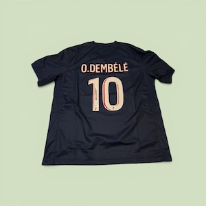 May include: Navy blue athletic jersey with the name "O. DEMBÉLÉ" and the number "10" in a cream colour. The jersey has a crew neck and short sleeves. The number "10" has a red outline.