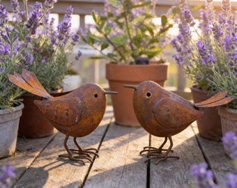 May include: Two rusty metal bird sculptures with outstretched wings stand on a wooden deck. The birds have a weathered appearance and are positioned in front of potted lavender plants. The scene is bathed in warm sunlight.
