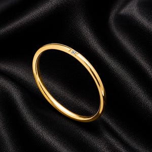 Op de afbeelding: Een dunne, gepolijste gouden ring met een enkele, kleine, heldere, rondgeslepen edelsteen die verzonken in het metaal is gezet. De ring heeft een glad, afgerond profiel en een heldere, reflecterende metalen afwerking met een strak ontwerp.