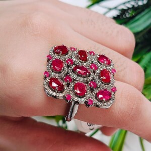 Puede incluir: Un anillo grande de forma cuadrada con múltiples rubíes rojos vibrantes de corte ovalado dispuestos en cuadrícula. Los rubíes están rodeados por pequeñas piedras blancas engastadas en pavé sobre una banda metálica, creando un diseño denso.