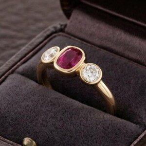 Op de afbeelding: Een gouden ring met een centrale kussengeslepen rode robijn, geflankeerd door twee ronde, helderwitte diamanten. De stenen zijn gezet in een gladde zetting op een gepolijste gouden band, gepresenteerd in een donkergrijze, met fluweel beklede doos.
