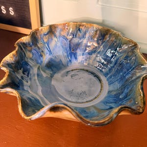 May include: A handmade ceramic bowl with a wavy rim. The bowl features a blend of blue, brown, and tan colors, creating a marbled effect. The interior is primarily blue, with the rim and outer edges showcasing brown and tan hues. The bowl is likely used for serving food or as a decorative piece.