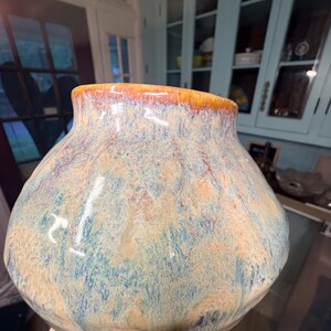 Könnte beinhalten: Eine handgefertigte Keramikvase mit breitem, rundem Körper und schmaler Öffnung. Die Vase hat eine Mischung aus Blau, Beige und Orange mit glänzendem Finish. Der Sockel ist weiß. Die Vase ist ein dekorativer Gegenstand.