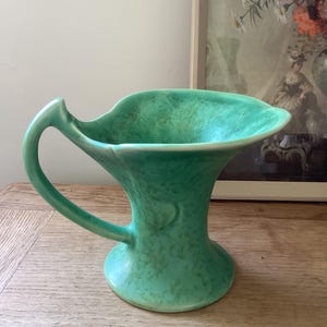 May include: A ceramic pitcher featuring a mottled mint green and light teal glaze. The vessel has a wide, flared rim with an asymmetrical, leaf-like shape, a curved handle on one side, and a circular base that tapers upwards into a narrow neck.