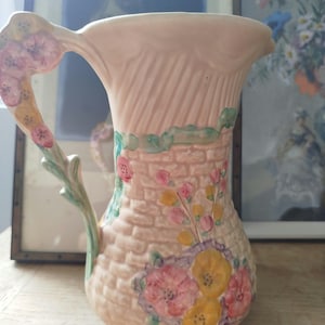 May include: A ceramic jug featuring a light peach-coloured body with a textured brick pattern. The jug is decorated with hand-painted pink and yellow flowers, green leaves, and a handle shaped like a vine with additional floral accents.