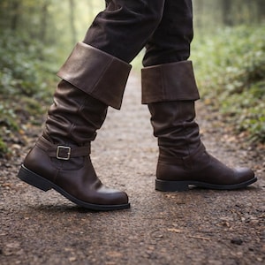 Op de afbeelding: Bruine leren kniehoge laarzen met een omgeslagen manchet en gesp. De laarzen worden gedragen met een donkerbruine broek en worden op een zandpad gelopen. De laarzen hebben een lichte glans en een klassiek design.