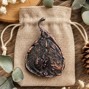 May include: A dark brown, pear-shaped pendant with a textured surface and a small, red gemstone is displayed on a beige burlap bag. The pendant has a curved metal loop at the top. The background includes wooden elements and greenery.