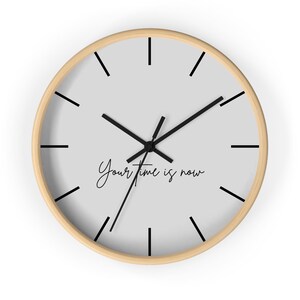Puede incluir: Reloj de pared redondo con esfera gris claro, manecillas y marcadores de hora negros. El reloj tiene un marco de color madera natural. Las palabras "Your time is now" están escritas en cursiva en la esfera.