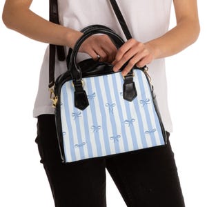 Puede incluir: Bolso de mano en blanco y negro con un estampado de rayas azul claro y blanco y detalles de lazos azul claro. El bolso tiene un asa superior y una correa para el hombro. El bolso lo sostiene una persona.