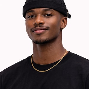 May include: A person wearing a black skull cap, a gold chain necklace, and a black t-shirt. The person is smiling and has a short beard and mustache. The background is plain white.