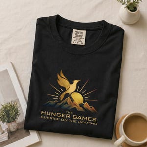 Pode incluir: Uma t-shirt preta de manga curta com um gráfico dourado de um pássaro a elevar-se sobre montanhas e um sol. O texto diz THE HUNGER GAMES: SUNRISE ON THE REAPING. A etiqueta da marca Comfort Colors no pescoço indica o tamanho large.