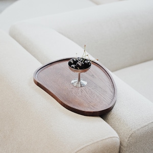 May include: A dark wooden serving tray with a unique shape rests on a cream-colored sofa. A small silver dish filled with dark cherries sits on the tray. The cherries have green stems. The sofa has a textured fabric.