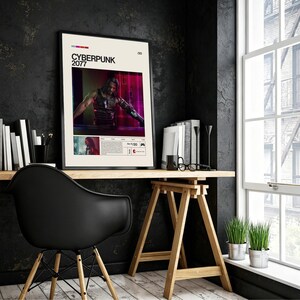 May include: A framed poster featuring the text "CYBERPUNK 2077" with an image of a character from the game. The poster is displayed on a wooden desk with books, a black chair, and a window in the background.