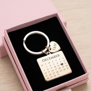 May include: A gold-colored keychain with a calendar for December, presented in a pink gift box. The keychain features a heart-shaped charm with "T&M" and a rectangular tag with the calendar dates. The box is open, revealing a black interior.