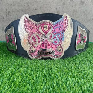 May include: A black wrestling championship belt with a large, ornate silver and pink center plate. The plate features the word "CHAMPION" and the letters "D.O.S." in a stylized font. The belt has two side plates with a pink and silver design.