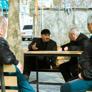 May include: A group of men are seated around a wooden table outdoors. The men are dressed in dark clothing, and one is wearing a black cap. A white car is visible in the background.