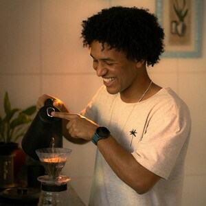 May include: A person pouring water from a black electric kettle into a glass pour-over coffee maker. The person is wearing a light grey t-shirt with a small palm tree graphic. A teal smartwatch is on the person's wrist.