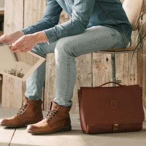 May include: A brown leather briefcase with a handle and buckle closure sits next to a person wearing brown boots and blue jeans. The person is sitting on a metal chair and holding a magazine. The background is a wooden wall.