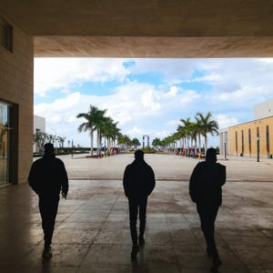 May include: Three silhouetted figures walk towards a bright outdoor scene. The scene is framed by a concrete structure, leading to a pathway lined with palm trees and colorful decorations. The sky is blue with scattered clouds.