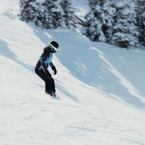 Peut inclure: Un snowboardeur dévale une pente enneigée. Il porte un casque noir, une veste bleue et noire, un pantalon noir et des gants noirs. L'arrière-plan montre des arbres enneigés et un ciel clair, typique d'une scène hivernale.