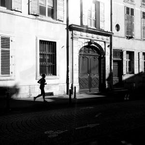 May include: Black and white photograph of a person running on a cobblestone street in front of a building with ornate doors and windows. The image is in sharp focus, with strong shadows and highlights.