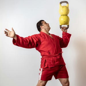 May include: A person in a red martial arts uniform balances two yellow kettlebells. The kettlebells are stacked on top of each other. The person is looking up with one arm outstretched.